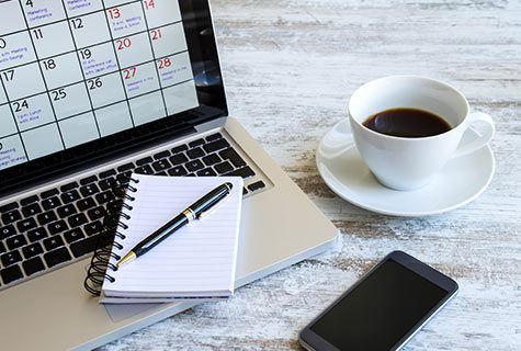A black smartphone resting on a white wooden table next to a cup of espresso and a laptop open to calendar with a white pad and pen resting on it.