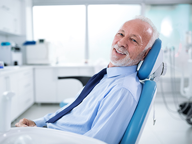 An elder man smiles in a blue shirt and tie as he sits in a teal dental chair.