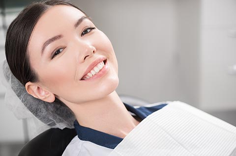 A smiling woman sitting in a dental chair and a cloth draped around her front in a dental room.